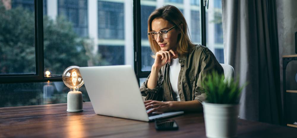 Frau mit Brille arbeitet an einem Laptop an einem Holztisch vor einem großen Fenster